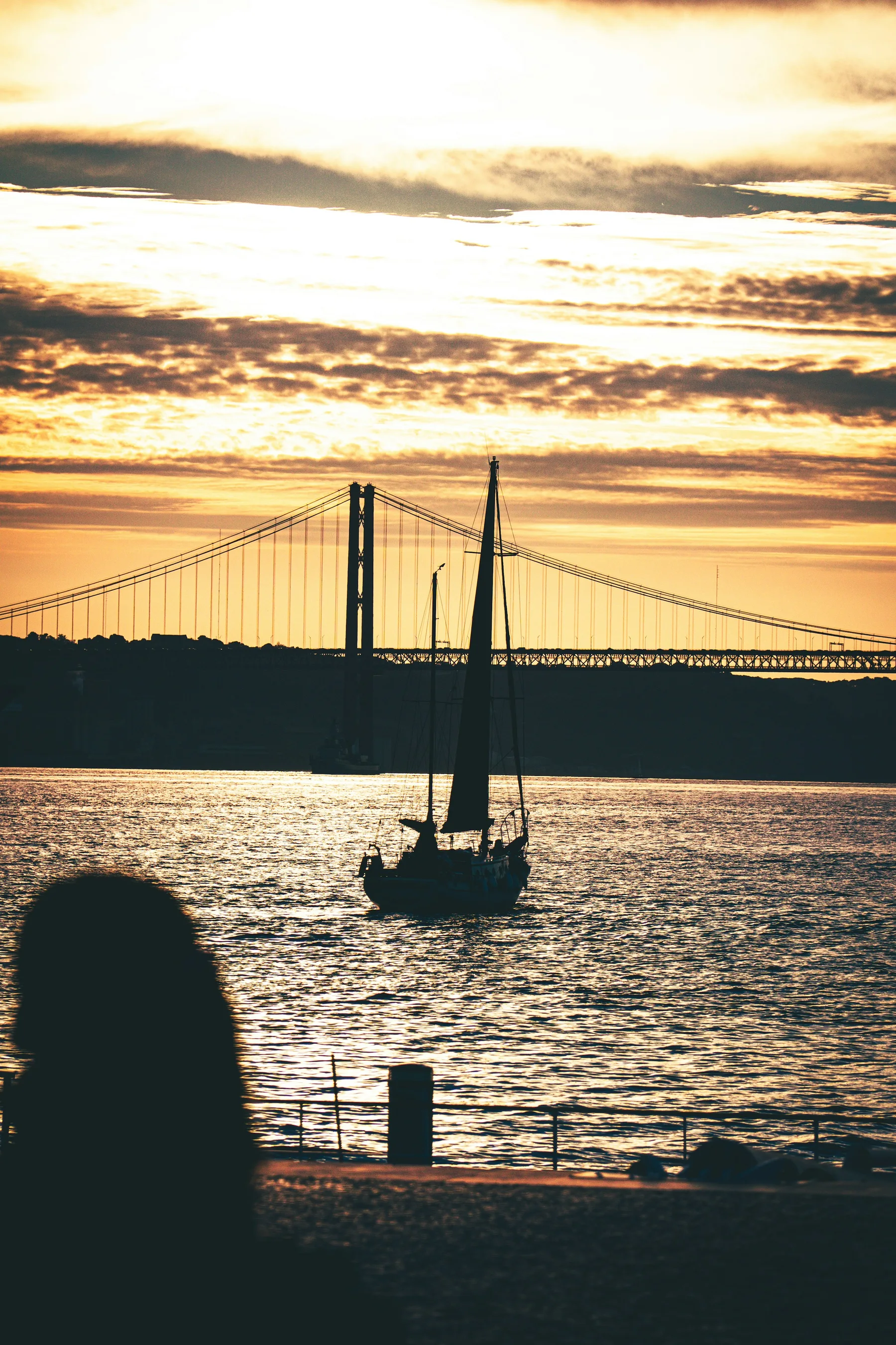 A sailboat silhouetted against Lisbon’s 25 de Abril Bridge at sunset.